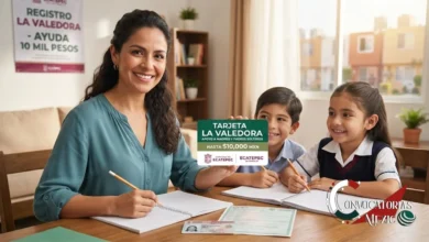 Una madre mexicana sonriente en su hogar en Ecatepec sostiene la tarjeta física 'La Valedora' de $10,000 MXN para apoyo a madres y padres solteros, mientras sus dos hijos hacen tareas. Sobre la mesa hay documentos de identidad (INE y CURP) y al fondo se ve un cartel oficial del programa.